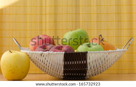 Closeup of a yellow apple next to a white basket full of red, yellow, green apples on a wooden surface in front of the yellow bamboo mat background