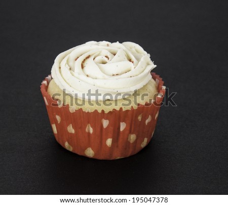 Vanilla cupcake with vanilla icing and red sugar sprinkles in a red liner with white hearts against a black background