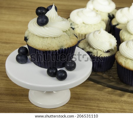 Cream cheese frosted blueberry muffin in dark blue paper with loose blueberries on a white pedestal against a plate of additional muffins and a brown wood background