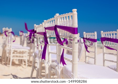 White wedding chairs decorated with purple bows on beach