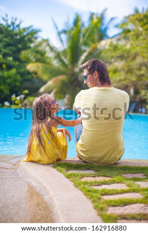 Back view of a young father and his cute daughter sitting by the swimming pool