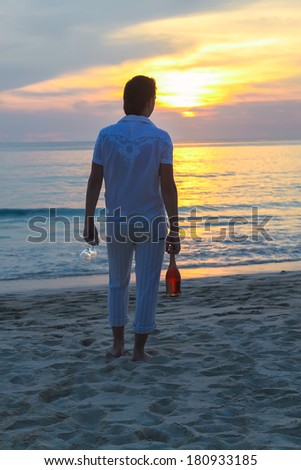 Young man with a bottle of wine and glasses on the beach at sunset