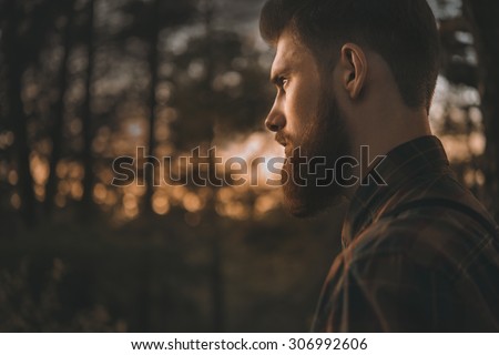 Brutal bearded man confidently looking forward. Standing alone in forest outdoor with sunset nature on background