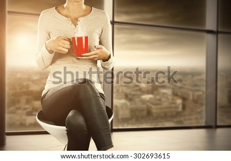 woman on white chair with red mug of hot drink and window with morning sun and city