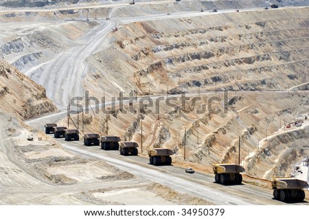 A long line of monster dump trucks drives down into an open pit mine to pick up their next load