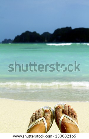 Vacations by the sea / Female feet wearing white flip-flops with a white sand beach and azure sea background, Phi Phi island, Thailand