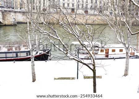 Tranquil winter scene in Paris : covered with snow river side of the Seine river with it\'s boats and parisian buildings visible on the other side.