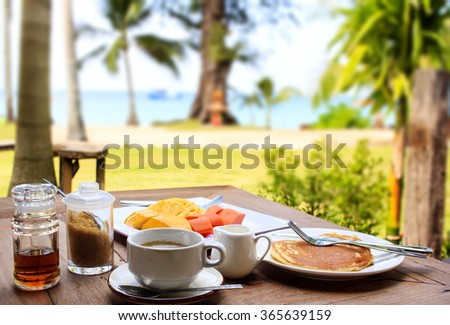Glorious early morning breakfast at the beach resort in Thailand : freshly brewed black coffee, pancake, maple syrup, fresh fruits. Tropical vegetation and the sea visible on the background.