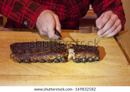 A man wearing a flannel shirt eating a rack of ribs off a wooden cutting board.