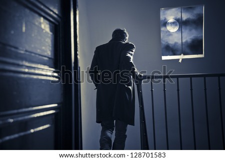 A man seen from behind is going down the staircase in an old parisian building at night. Spooky moon and clouds in the window.