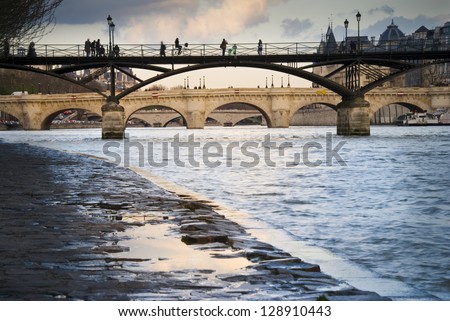 Passerelle des Arts over River Seine in Paris, France. Silhouettes of people walking on it. Pont Neuf (the oldest bridge in Paris) in the distance.