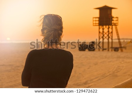 Young woman seen from behind gazing at the sunset on Conil de la Frontera beach, in Andalusia, Spain. Lifeguard watchtower in the background.