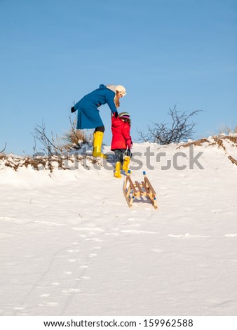 Adult woman helps small girl pulling a wooden sledge to the top of a snow covered dune