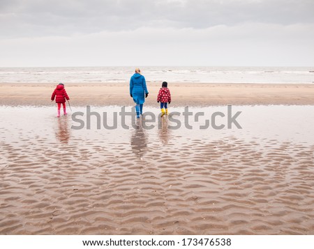 woman with small boy and girl in winter clothing and rubber boots paddling in a tidal pool on a winter beach looking out to sea