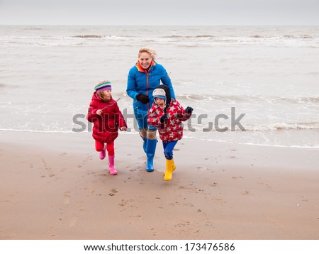 woman with small boy and girl in winter clothing and rubber boots playing on a winter beach running away from waves