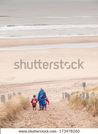 woman with small boy and girl in winter clothing and rubber boots walking down to a winter beach along a dune path
