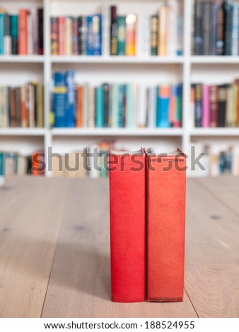 Aged copies of old red bound books standing on a wooden table with a book shelf full of books in soft focus behind