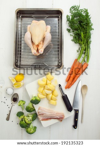 ingredients for a roast chicken dinner with whole chicken, vegetables and utensils on a rustic white table top