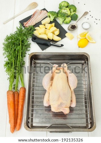 raw whole chicken on a roasting tray with vegetables and utensils on a white rustic table top