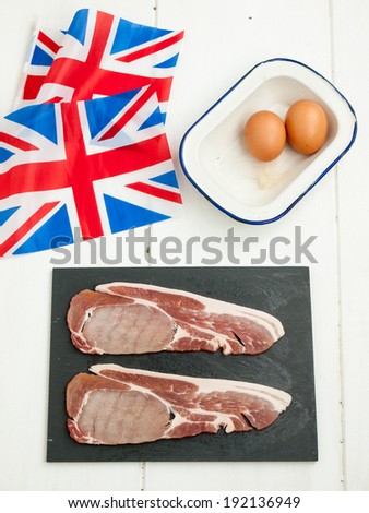 two raw thick cut back bacon rashers with eggs on a rustic white table top with a british flags