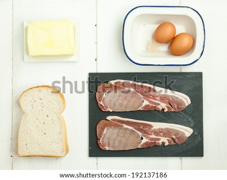 raw thick cut back bacon rashers with eggs, sliced white bread and butter on a rustic white table top