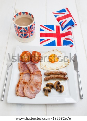full fried english breakfast on a square white plate on a rustic white table top with knife and fork and british flags and cup of tea