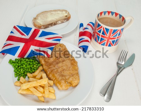 a plate of british fish and chips with peas, a slice of bread and butter, a mug of tea in a union jack mug and british flags