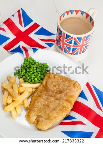 closeup of a plate of british  fish chips and peas and a mug of tea in a union jack mug and british flags on a rustic white table top