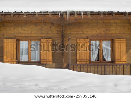 shuttered cabin windows behind a snow drift below a snow covered roof with icicles