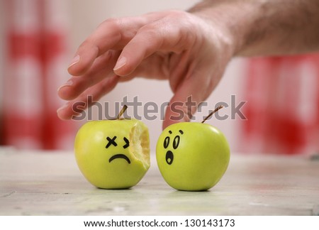 One bitten apple and another one with comic painted face looking worried about the hand from above