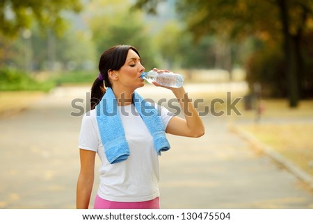 Beautiful young sporty woman with towel around neck drinking water after fitness exercise