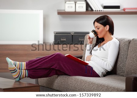 Young Woman Lounging In Living Room On Couch Reading Book And Drinking Coffee