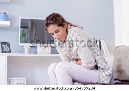 Young woman sitting on the bed with hard stomach pain.\
Shallow depth of field.