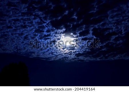 Full moon with indigo hues glowing in night sky illuminating clouds in wide angle perspective/Full moon glowing in night sky illuminating cloud cover