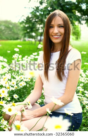 The young beautiful woman sits on a green field on which camomiles grow.