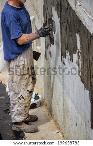 Exterior basement wall waterproofing, worker installing waterproofing tar sealer. Selective focus and motion blur.
