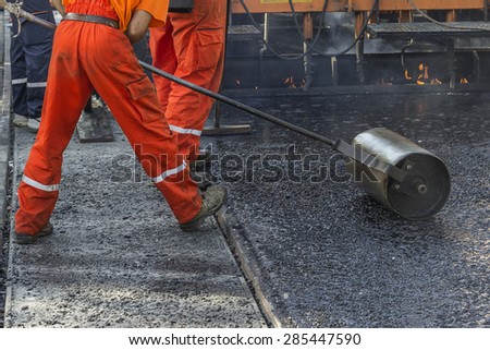 Worker pushing hand roller for mastic asphalt paving, making asphalt with coated chippings. Selective focus and shallow dof, some motion blur present.