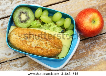 Packed lunch box containing brown cheese roll, grapes, kiwi fruit and red apple
