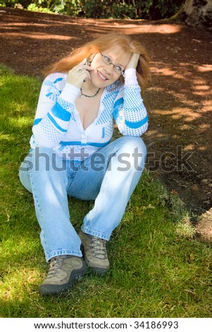Beautiful Redhead Woman day dreaming and talking on Cell Phone sitting on green grass under the shadows of a tree. She\'s wearing blue jeans and is wearing glasses and a necklace.