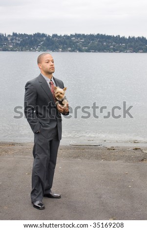 Ethnic Business Man and Yorkshire Terrier Dog are standing next to the lake. He is dressed in a suit and tie and seems to be contemplating something.