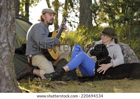 Senior couple enjoying some music outside their tent in evening light. Man plays guitar and sings. They have their pet curly coated retriever with them