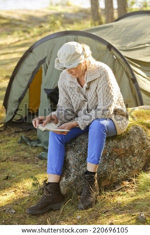 Senior woman sits outside her tent reading a book. There\'s some digital filter flare