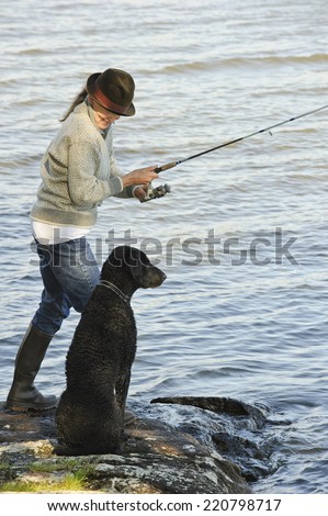 Senior woman fishing from the lake shore. Her curly haired retriever pet sits beside her