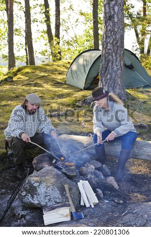 Senior couple grill sausages over camp fire using sticks