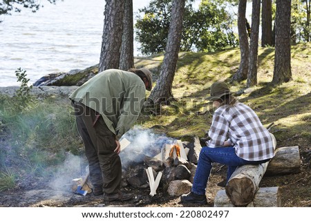 Senior couple light a camp fire. They are in a forest beside a lake