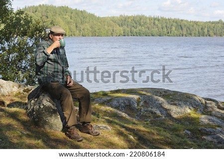Senior man sits on a rock by a lake surrounded by forests. He\'s drinking from a coffee or tea mug.