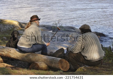 Senior couple sitting by camp fire near lake. Curly haired retriever pet sits beside them. The woman has a sausage on a stick