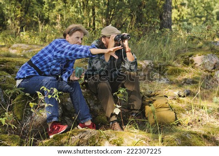 Senior couple have a tea or coffee break in the forest while hiking. They use  binoculars to check out the landscape. Focus is on the woman