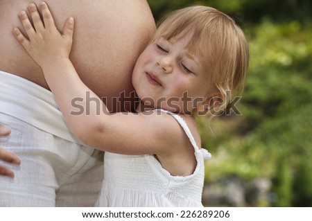 3 year old blond girl dressed in white listening to her pregnant mother\'s bared stomach - mother also dressed in white. Located in a summer garden with green out of focus background.