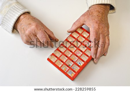 Hands of an eighty eight year old man open a pill organizer box. The box is filled with a variety of pills, and is divided into four times a day, seven days a week